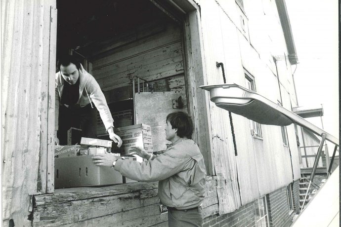Photo, black and white: Two men pass boxes from an open warehouse door to the trunk of a car. 