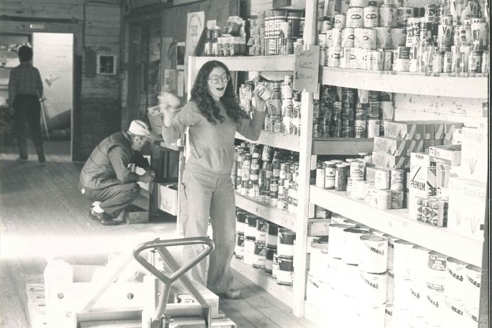 Photo, black and white: A young woman with large glasses places cans on a wooden shelving unit.