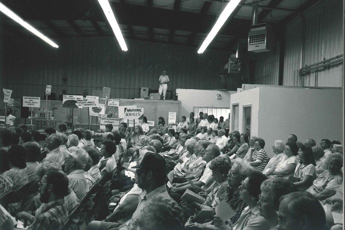Photo, black and white: An event to dedicate the new Hatfield warehouse - a crowd sits seated in a large open interior space. 