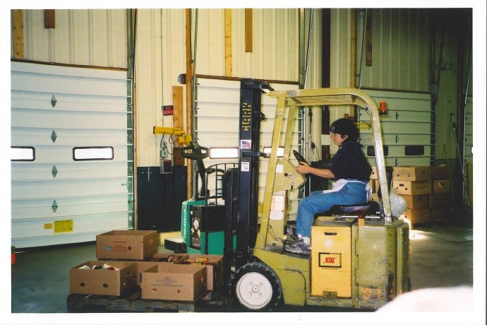Image, color: A woman drives a forklift, lifting a set of produce boxes in front of warehouse garage doors. 