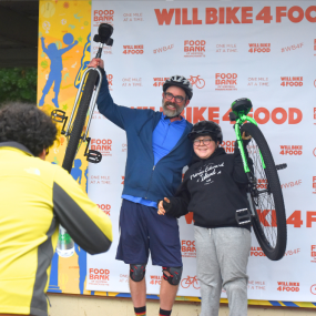 Two cyclists pose with their unicycles in front of a Will Bike 4 Food background.