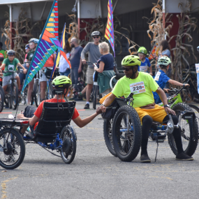 Two cyclists on accessible recumbent bikes shake hands at Will Bike 4 Food.