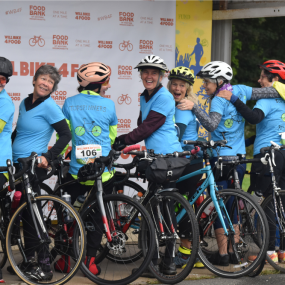 A group of smiling cyclists pose in front of a Will Bike 4 Food background.