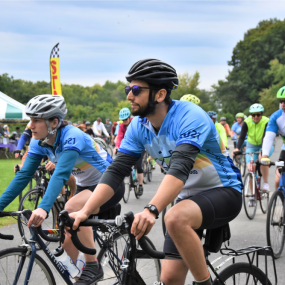 Two cyclists pass in front of the camera wearing Will Bike 4 Food jerseys, with others in the background.