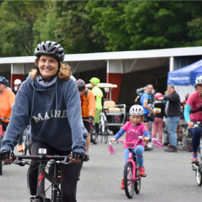 A smiling cyclist rides towards the camera, with others visible in the background.