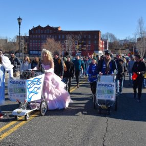Monte Belmonte, Dressed as Glinda and accompanied by Dorothy, pushes a shopping cart down the center of a city street followed by a crowd of other marchers.