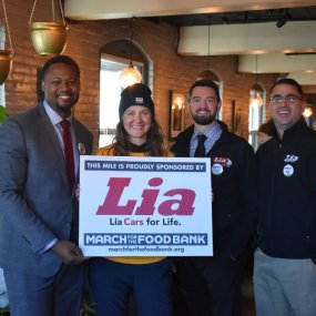 A group of employees from March Sponsor Lia Toyota pose with a sign announcing their sponsorship and a Food Bank staff member.