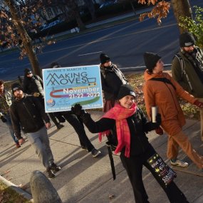 A person walking down a city sidewalk, surrounded by other marchers, holds a sign for "Monte's March" up to the camera.