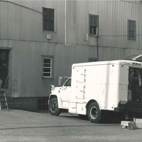 An archival print photo of a white refrigerated truck in front of the open door of a loading dock.