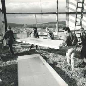 An archival print photo of a team carrying large wall panels within the open steel frame of a building under construction.