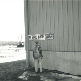 An archival print photo of a gentleman standing in front of a sign reading "The Food Bank, Coming Soon", with construction equipment in the background.