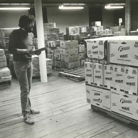 An archival print photo of a person documenting a palette of Wheat Snack Crackers on a small clipboard, with large palettes of other dry foods in the background.