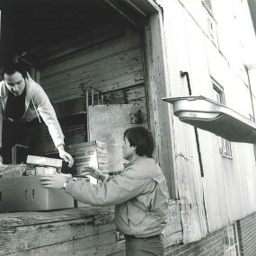 An archival print photo of two people passing cardboard boxes of food from the open trunk of a car through an open loading dock door.