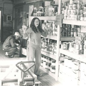 An archival print photo of a woman sorting cans on series of large wooden shelves.