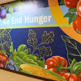 A colorful mural on the wall of a hallway at the Food Bank, featuring a variety of fruits and vegetables and reading "Together, We End Hunger".